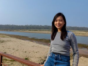 A photo of Jocelyne Sze, a young woman with medium length black hair. Behind her is a river and a forest. 
