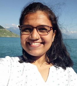 Photo of Manasi Mulay a young woman with long dark hair and glasses smiling in front of an ocean. 