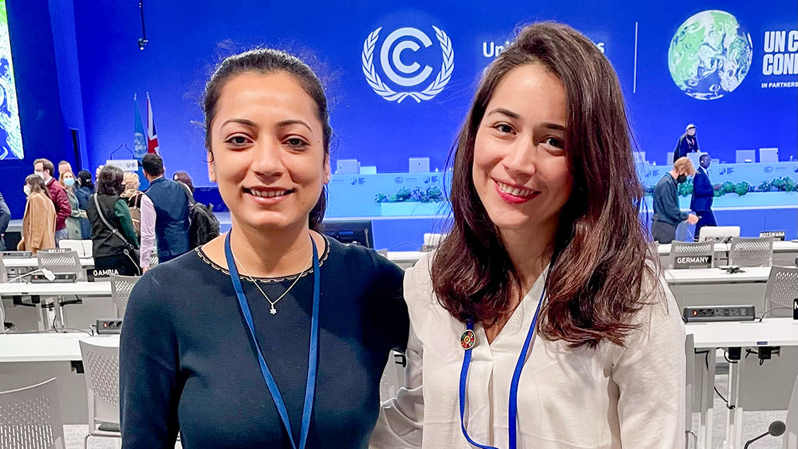 Grantham Scholar Reena Sayani stands with Paloma Ortega from the Grantham Institute in front of a COP26 banner. They are both young women with dark hair hair wearing COP lanyards and smiling.