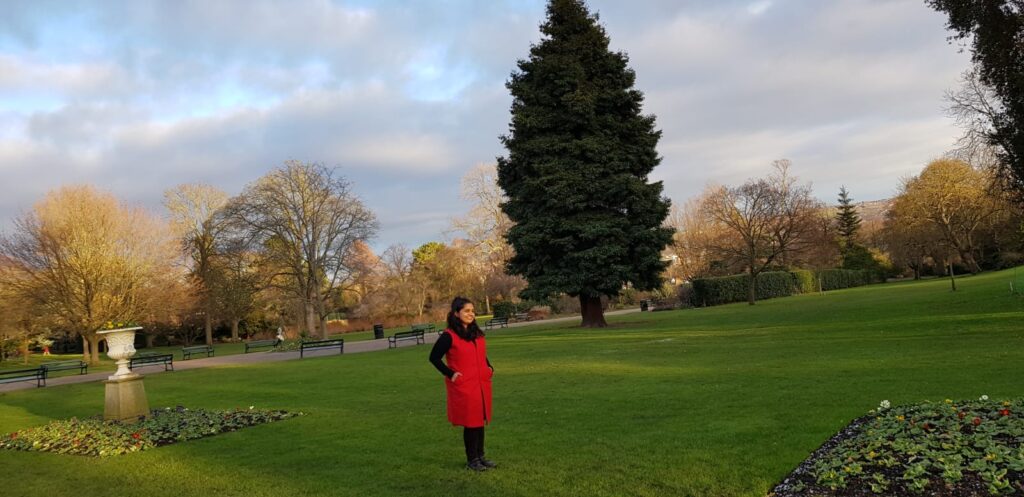Manasi Mulay - young Indian woman in an orange pinafore - stands in a botanical park in Sheffield. Behind her are trees, deciduous without leaver and one tall evergreen. She stands on a grass lawn and near her is a stone plant pot. 